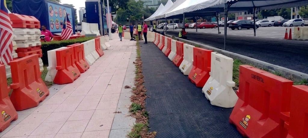 Red and white plastic road barriers from Cone Rental lining a pedestrian route at a public event in Putrajaya, with event tents and Malaysian flags behind. Event traffic management and road closure equipment rental in Malaysia.